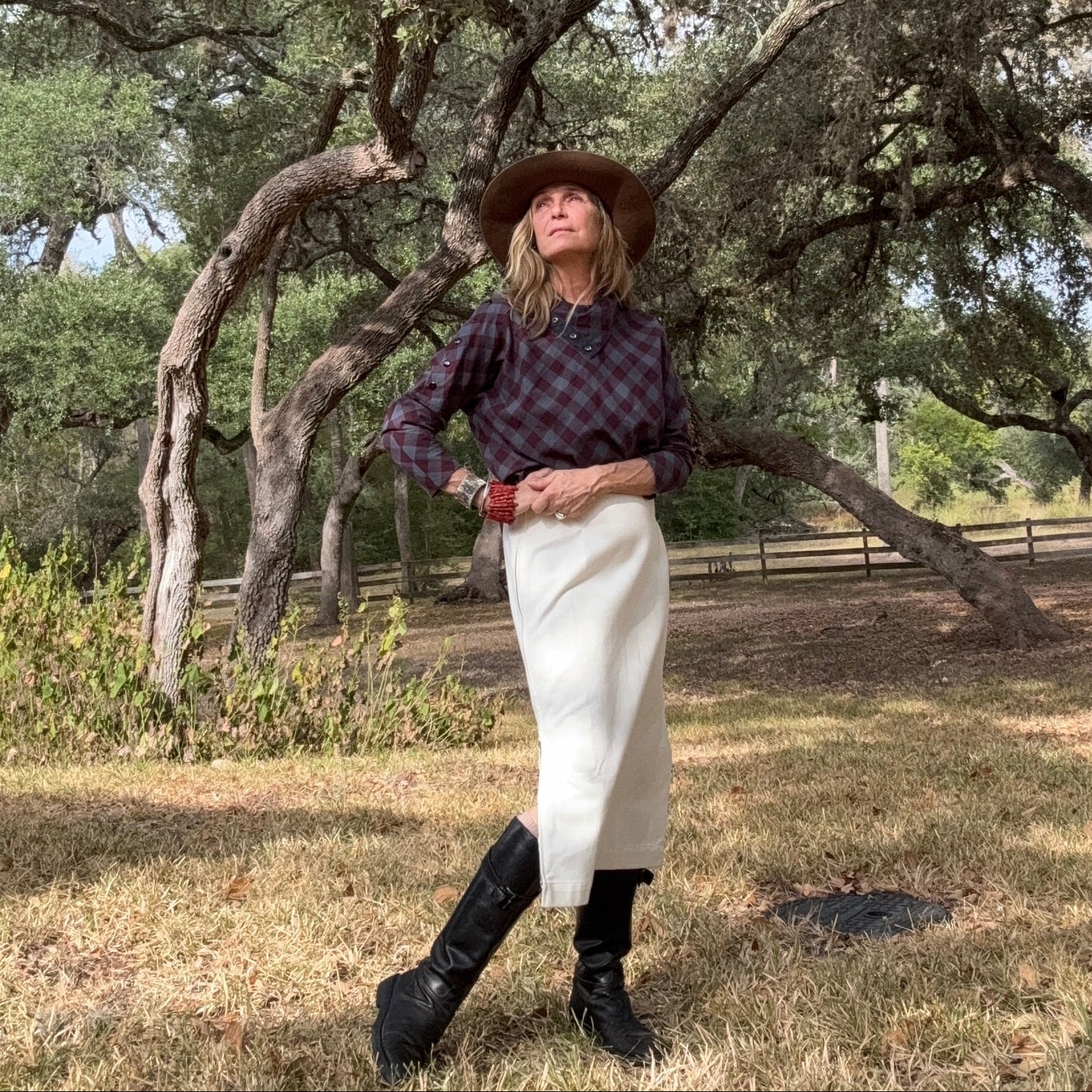 Person wearing a plaid shirt, white skirt, and cowboy hat standing in a natural setting with trees.