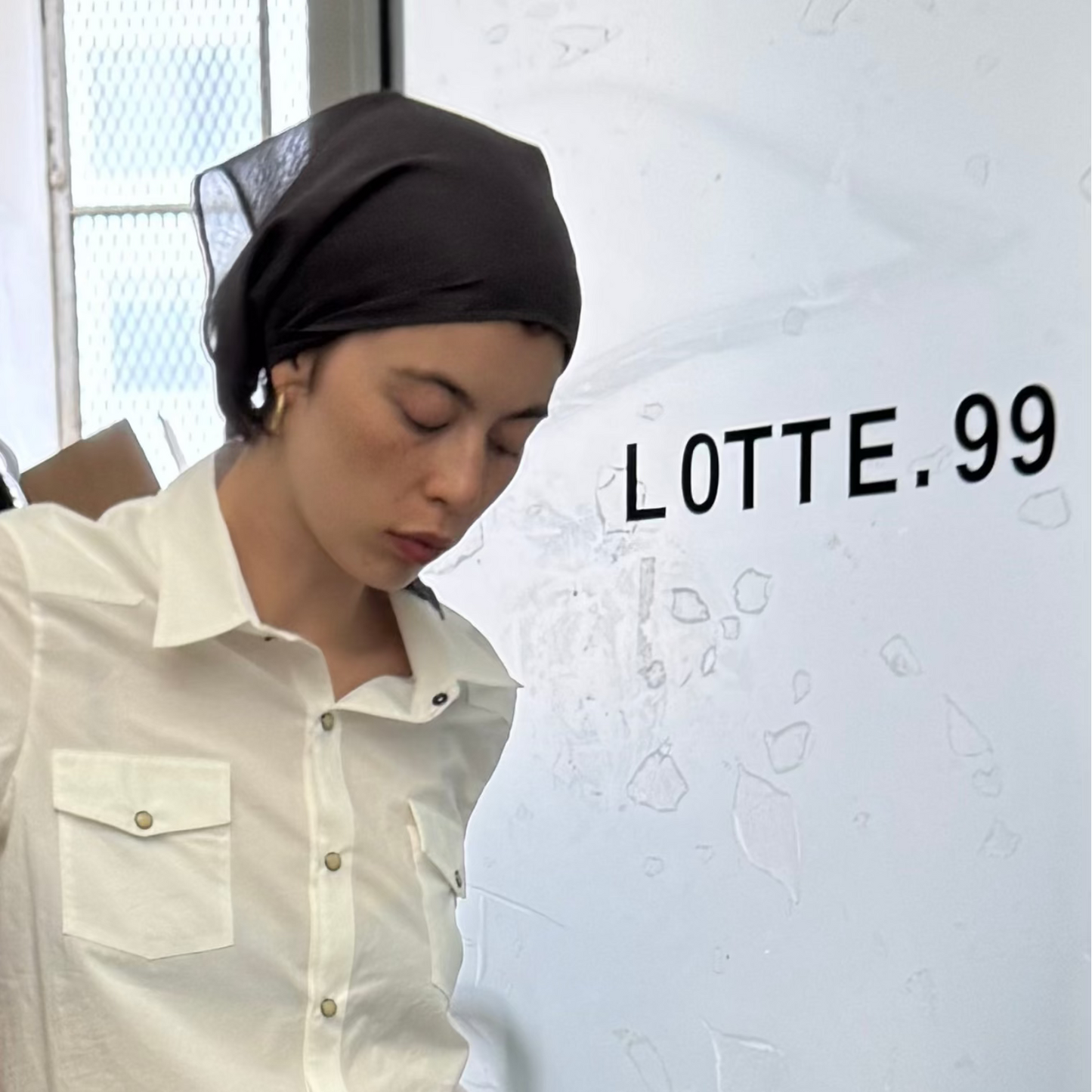 Person wearing a white shirt and headscarf in front of a clothing rack with 'LOTTE.99' branding.