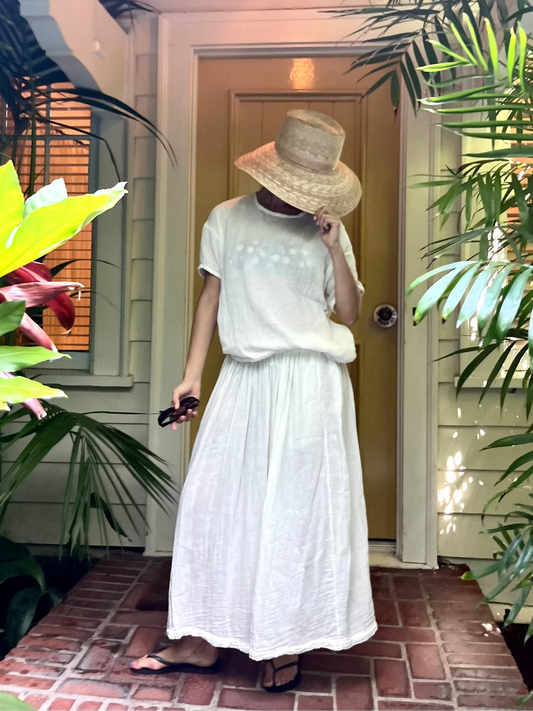 A person wearing a white short-sleeve tunic and matching long skirt stands on a brick porch surrounded by lush green plants, holding a straw hat and sunglasses.