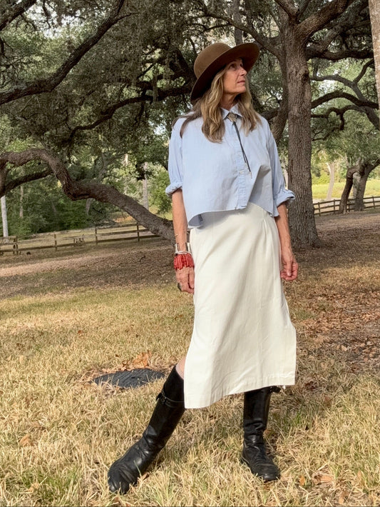 Woman in a blue shirt, white skirt, and brown hat standing in a park with trees and grass.
