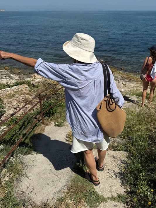 A person wearing a white bucket hat, a light blue button-up shirt, and white shorts stands on a rocky seaside path pointing toward the ocean, with a tan shoulder bag and another person in swimwear in the background.