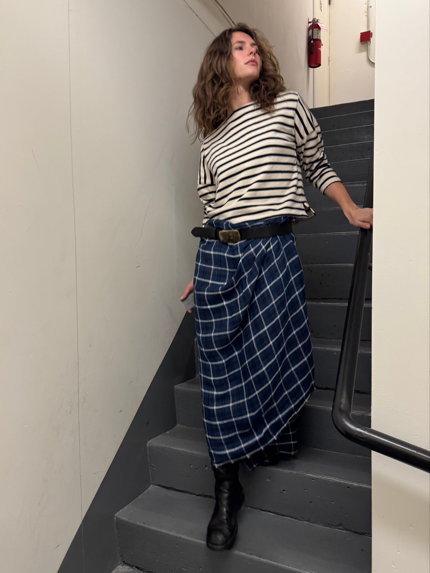 A woman stands on an indoor staircase wearing a black and white striped long-sleeve top, a navy blue and white plaid skirt, and black boots.