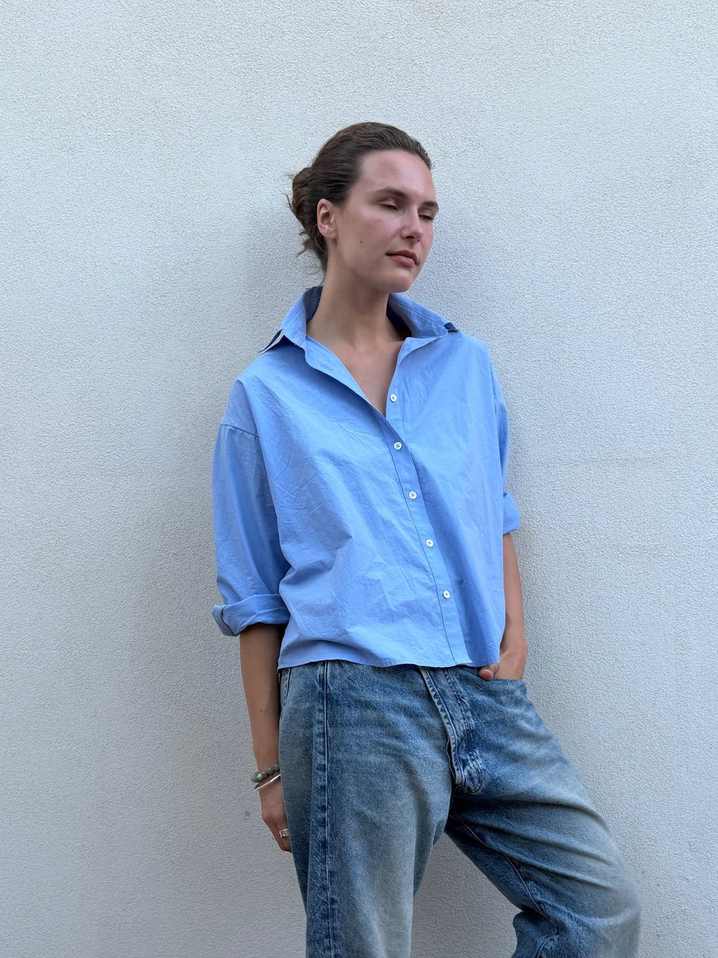 A woman stands against a light textured wall wearing a light blue button-up shirt, wide-leg blue jeans, and dark sandals.