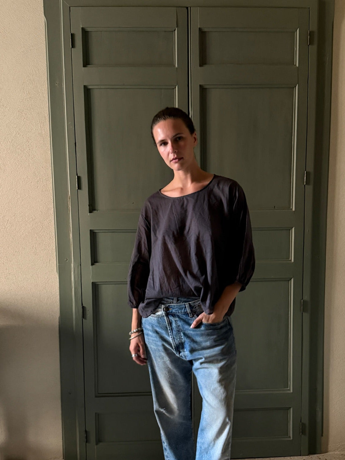 A woman stands in front of a large dark green paneled door wearing a dark purple long-sleeve top, light blue jeans, and sandals.