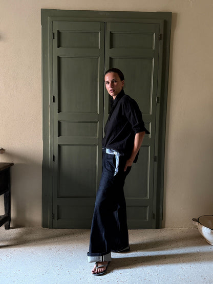 A woman stands in a room wearing a black long-sleeve shirt tied loosely at the waist with a light blue ribbon, dark wide-leg pants, and sandals. She is posed in front of a large dark green paneled door.
