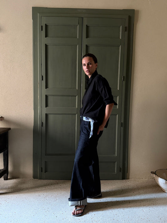A woman stands in a room wearing a black long-sleeve shirt tied loosely at the waist with a light blue ribbon, dark wide-leg pants, and sandals. She is posed in front of a large dark green paneled door.