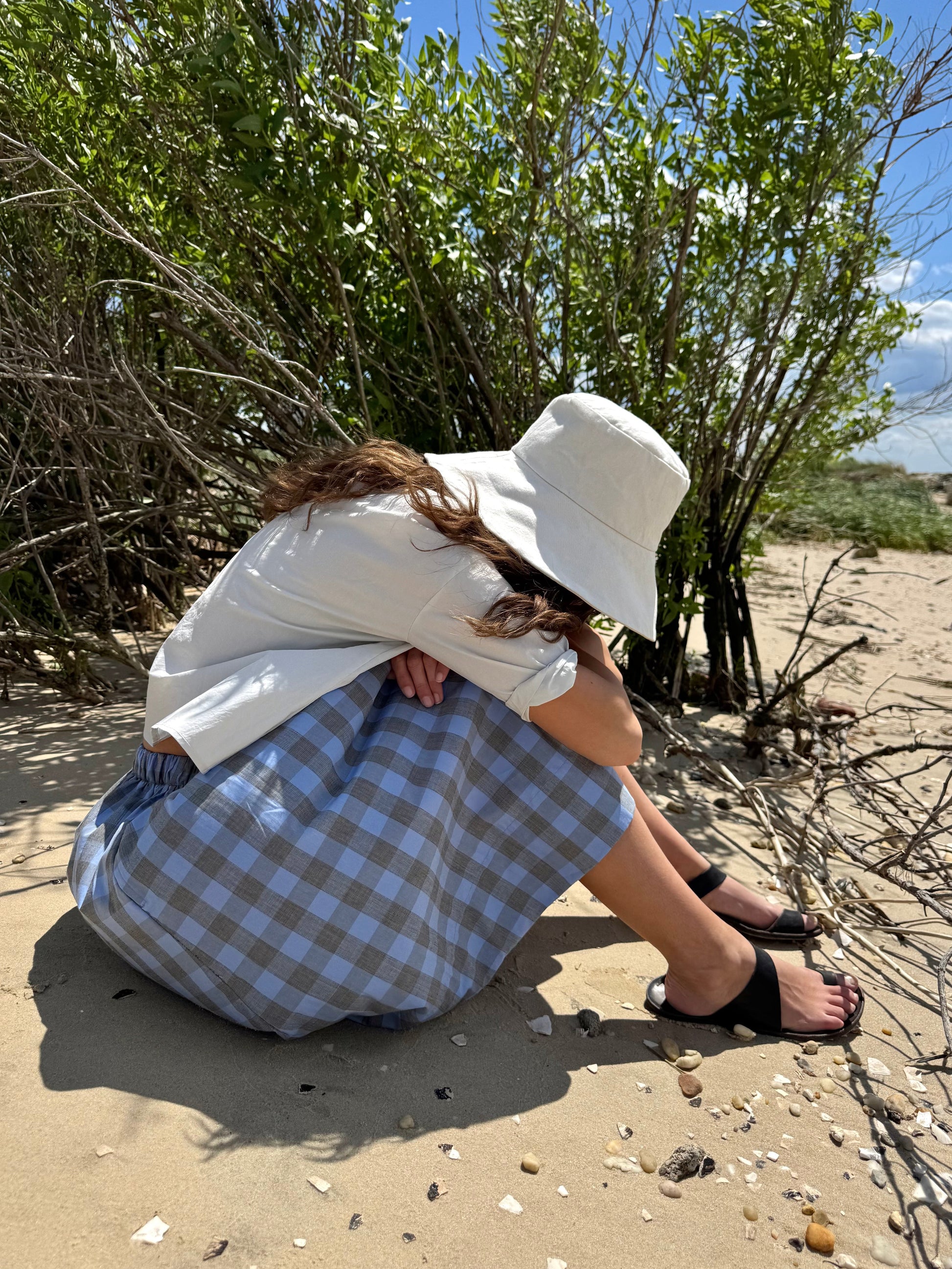 A person crouching on sandy ground near leafy shrubs under a blue sky, wearing a white bucket hat, white top, blue-and-white gingham skirt, and black sandals.
