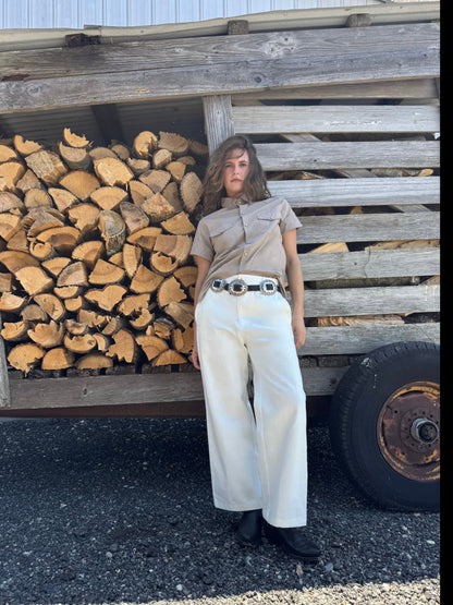 A woman stands outdoors in front of a rustic wooden structure filled with stacked firewood. She is wearing a beige short-sleeve collared shirt, off-white wide-leg pants with a black belt, and black boots.